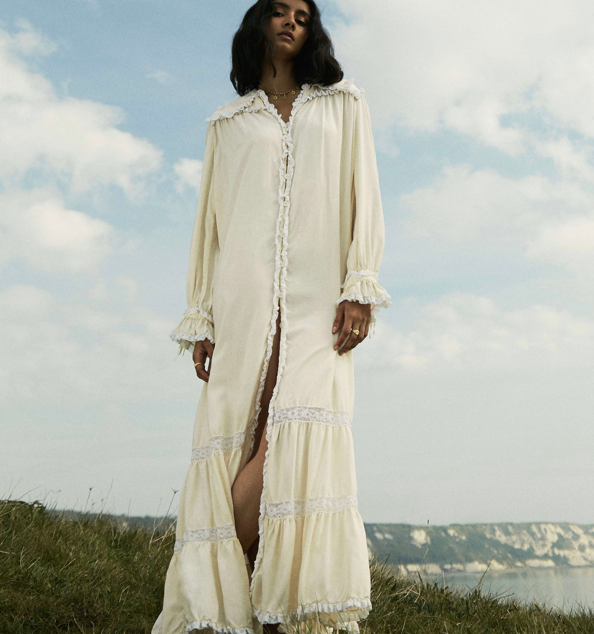 Woman wearing gold layered necklaces and a gold ring, standing in a grassy field against a cloudy sky.
