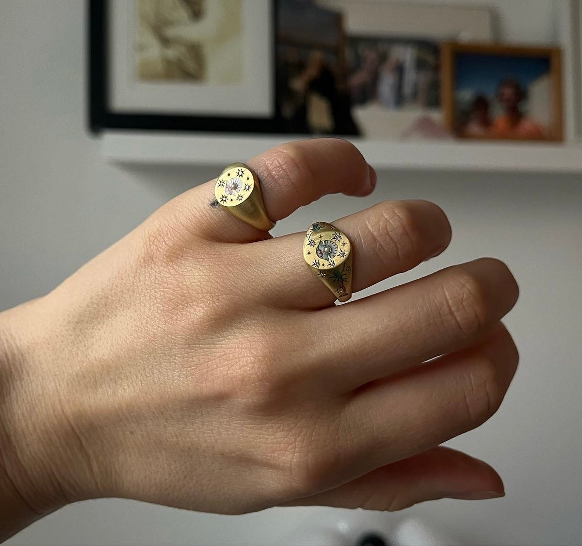 Woman wearing two gold rings featuring floral enamel motifs on a hand against a minimalist background.