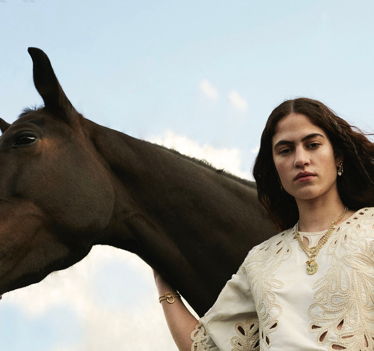 Woman wearing gold earrings and layered gold necklaces with a horse against a blue sky.