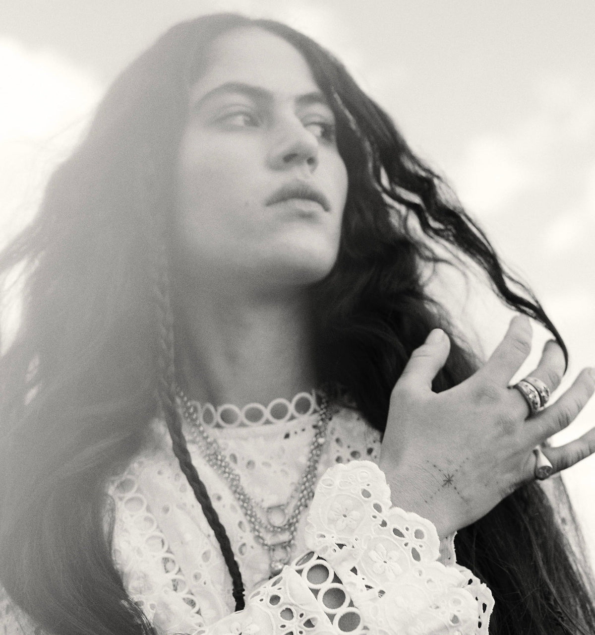 Woman wearing multiple gold necklaces and rings, with long hair and a white lace top against a cloudy background.