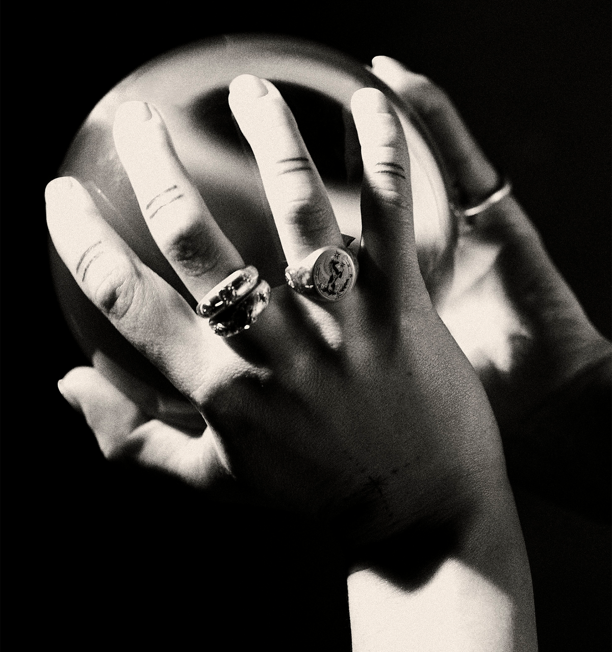 Woman wearing multiple gold rings, gently holding a crystal ball with both hands against a dark background.