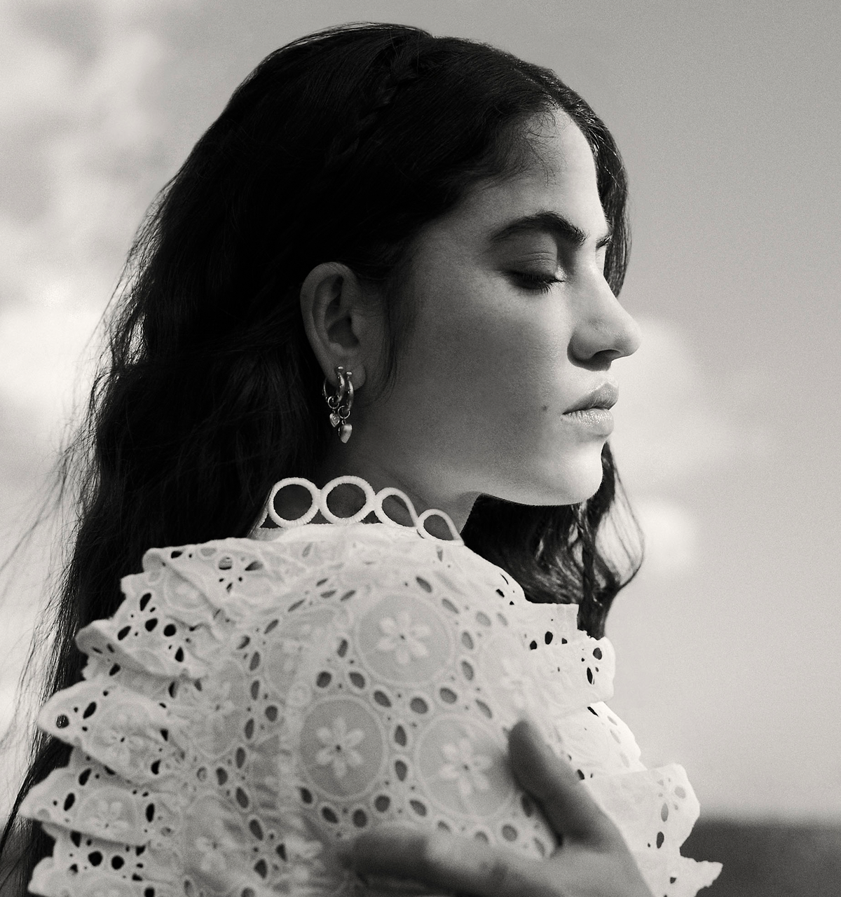 Woman wearing gold earrings against a blurred outdoor background, with an intricate white lace top.