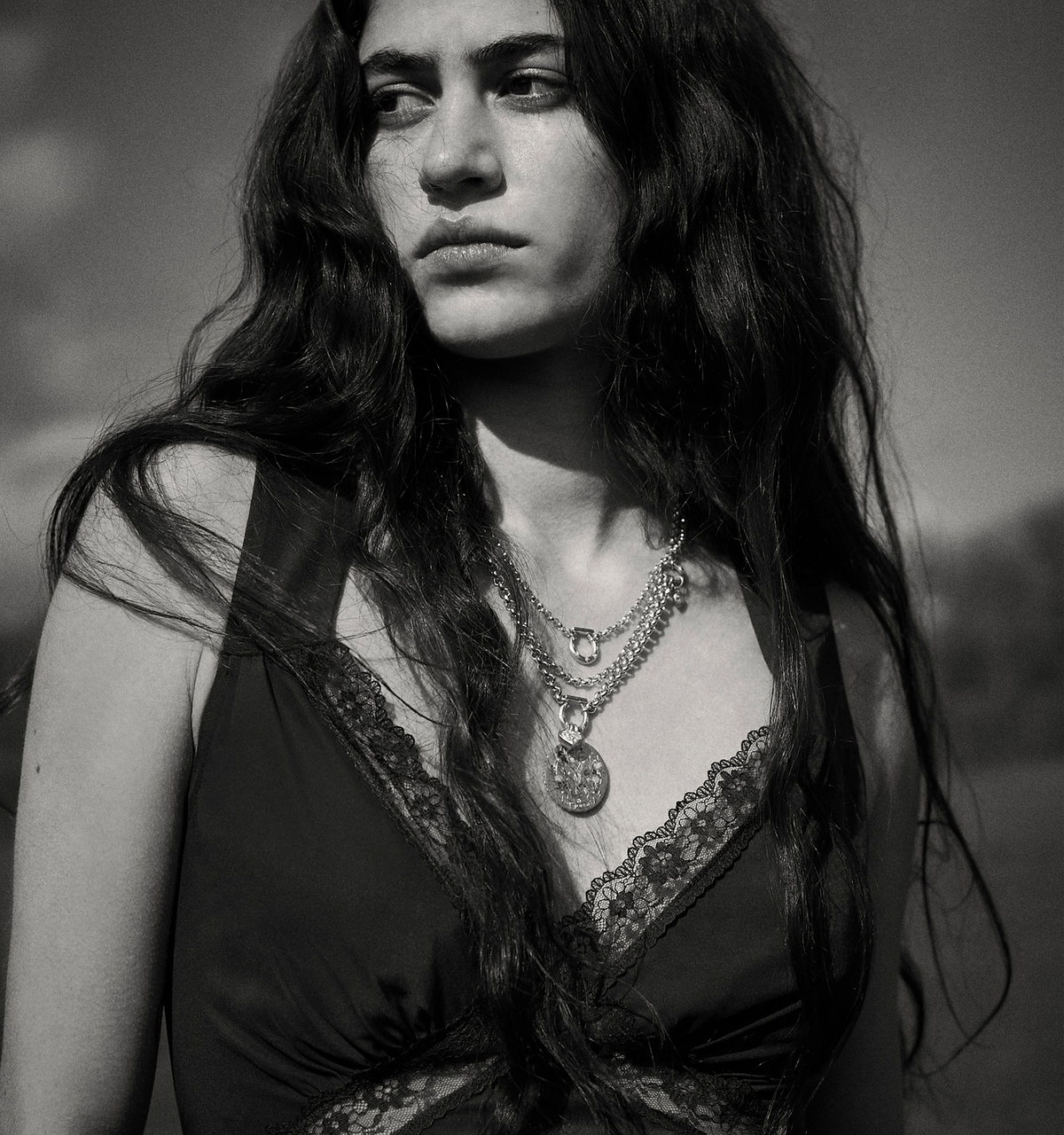 Woman wearing layered gold necklaces with a circular pendant, in a field setting, with long wavy hair.