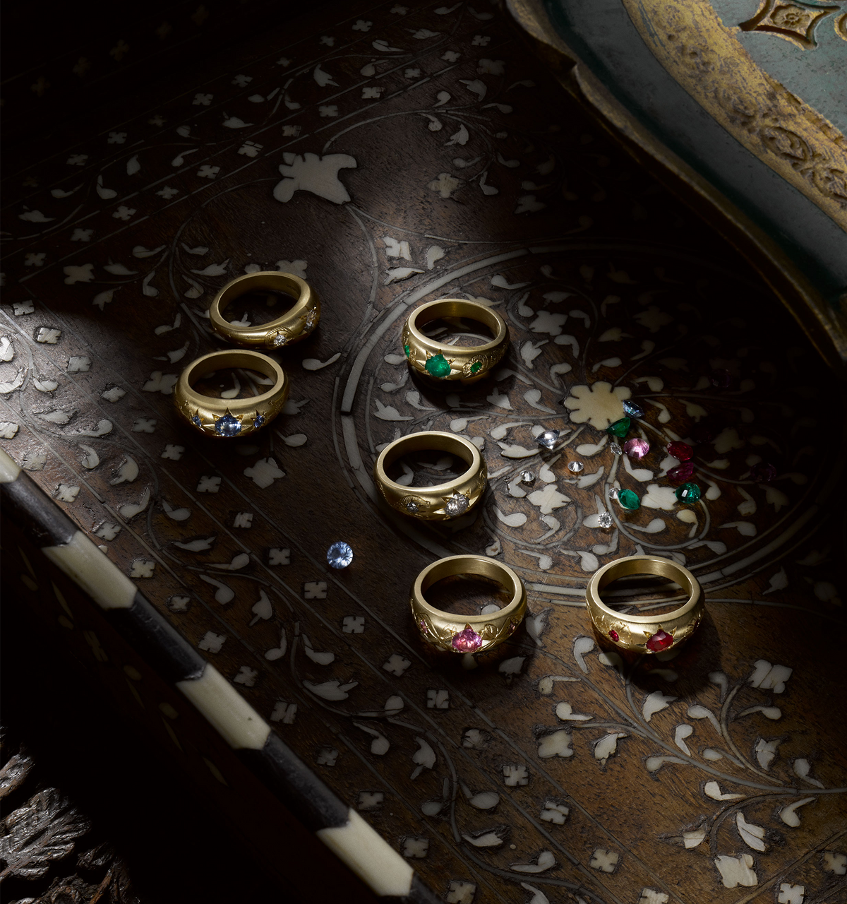 Gold rings adorned with emeralds, sapphires, and rubies on an intricately patterned wooden surface.